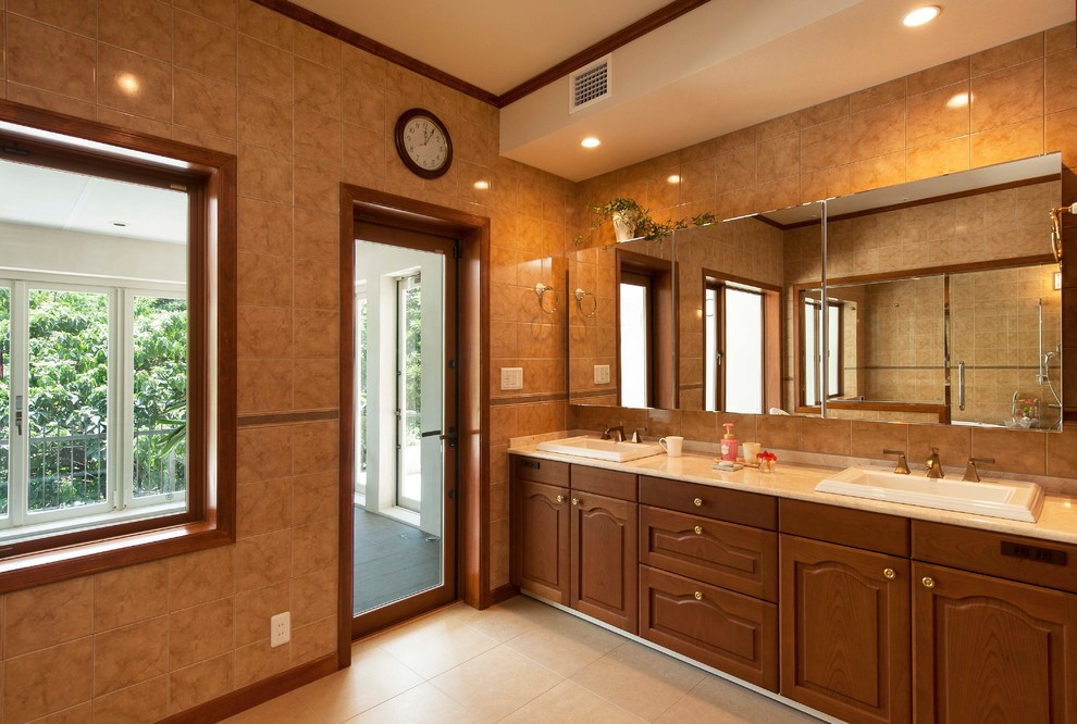 他の地域にある地中海スタイルのおしゃれな浴室 (落し込みパネル扉のキャビネット、中間色木目調キャビネット、茶色いタイル、茶色い壁、オーバーカウンターシンク、ベージュの床) の写真