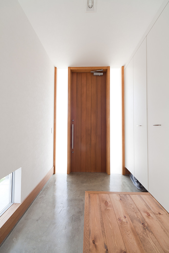 Cette image montre une entrée minimaliste avec un mur blanc, sol en béton ciré, une porte simple, une porte en bois brun et un sol gris.